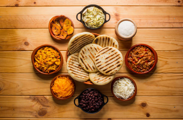 View from above of a wooden rustic table with several ingredients for cooking and filling arepas, typical Latin American food.  In the image we can see arepas, meat, chicken, black beans, cheeses, avocado, corn and some spices and vegetables